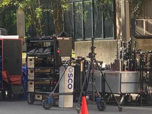 Carts loaded with cases and sandbags sit beside stacked light stands and grip equipment during an outdoor film shoot.