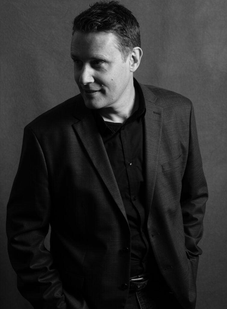 Studio portrait in black and white of a man wearing a dark blazer and shirt, hands in pockets, looking off-camera.