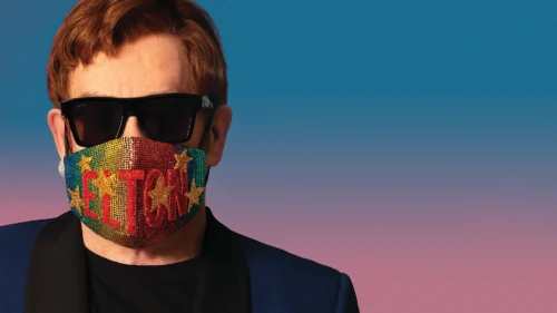 Close-up of a person wearing black sunglasses and a colorful rhinestone face mask that reads “ELTON” against a blue-to-pink gradient background.