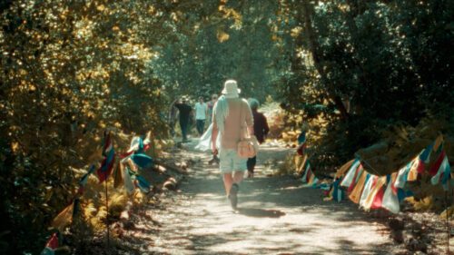 People walking along a sunlit woodland path lined with colorful prayer flags.