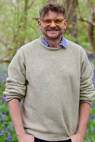 Smiling person wearing round yellow glasses and a beige sweater, standing outdoors in a green woodland.