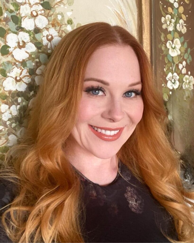 Close-up portrait of a woman with long wavy red hair and makeup, smiling against a decorative floral backdrop.