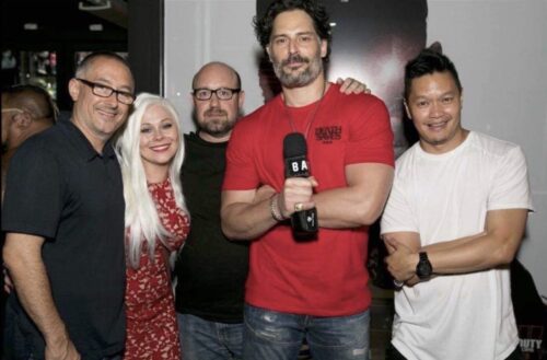 Five people smiling for a photo indoors; one holds a microphone while others stand close together.
