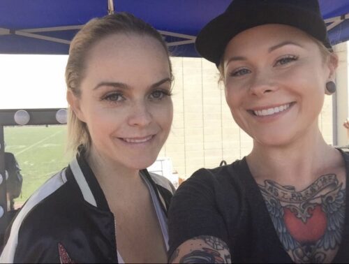 Two women pose for a selfie at an outdoor event; one wears a black cap and has visible chest tattoos.