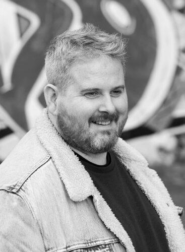 Black-and-white portrait of a smiling person with short hair and a beard wearing a shearling-lined denim jacket, with graffiti in the background.