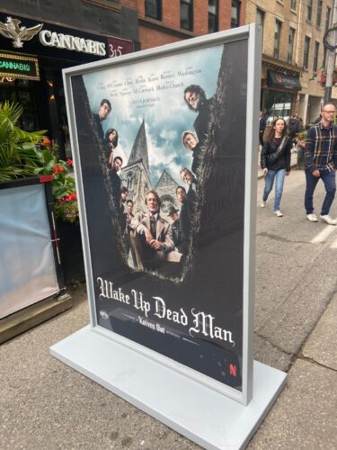 Large freestanding movie poster for “Wake Up Dead Man” on a downtown sidewalk, with pedestrians walking past.